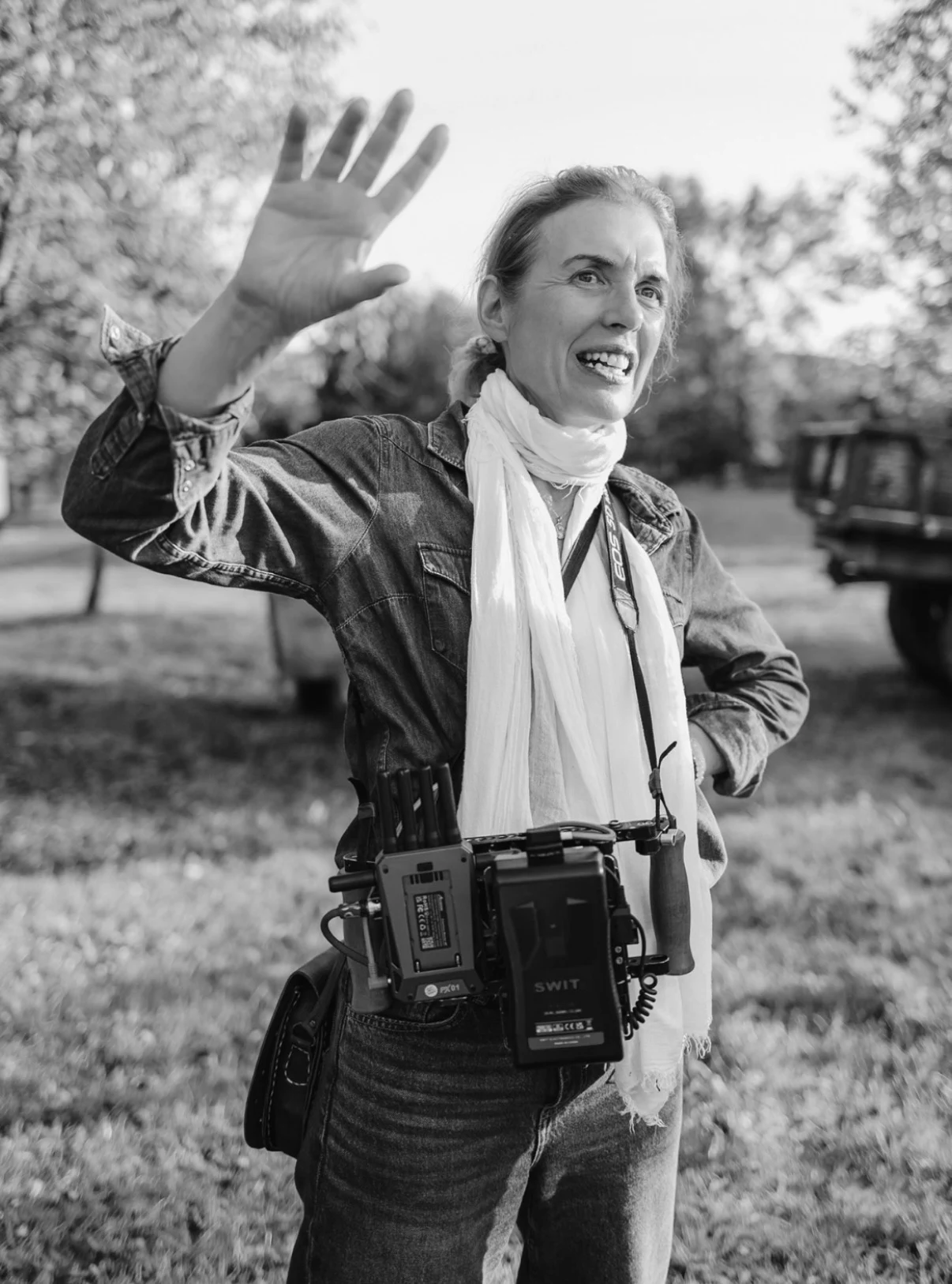 Diane Bell smiling and raising a hand on set, with a camera monitor around her neck